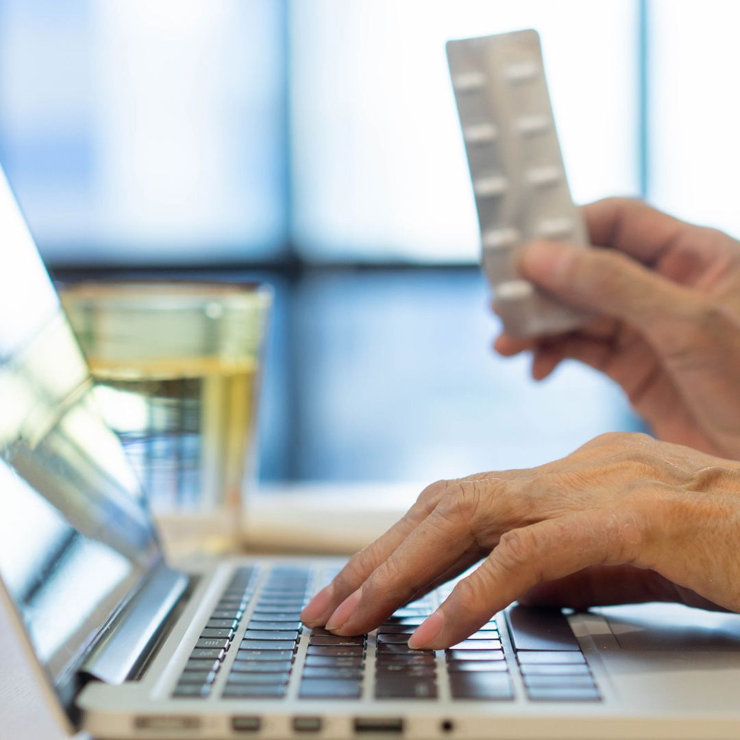 A person is typing on a laptop computer while holding a blister pack of pills.
