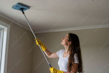 Woman in yellow gloves using a ceiling duster, cleaning a white ceiling in a room.
