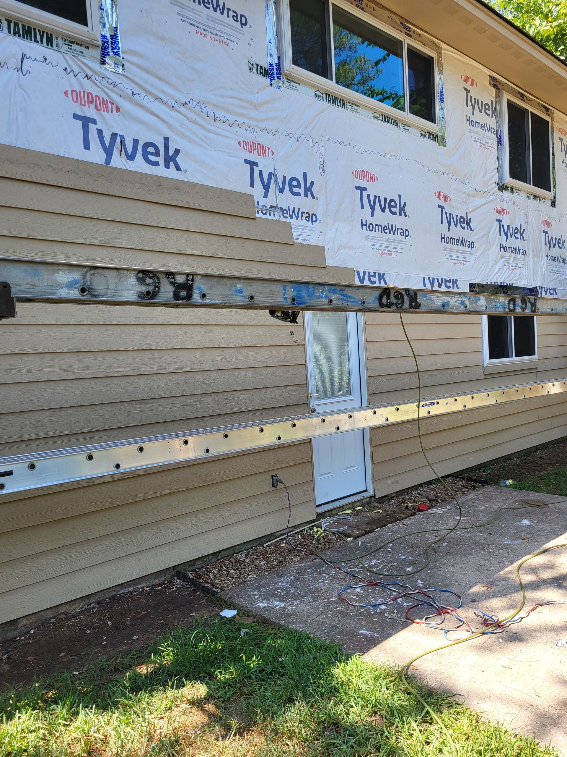 Person painting siding of a house, standing on an orange ladder. Blue sky in the background.