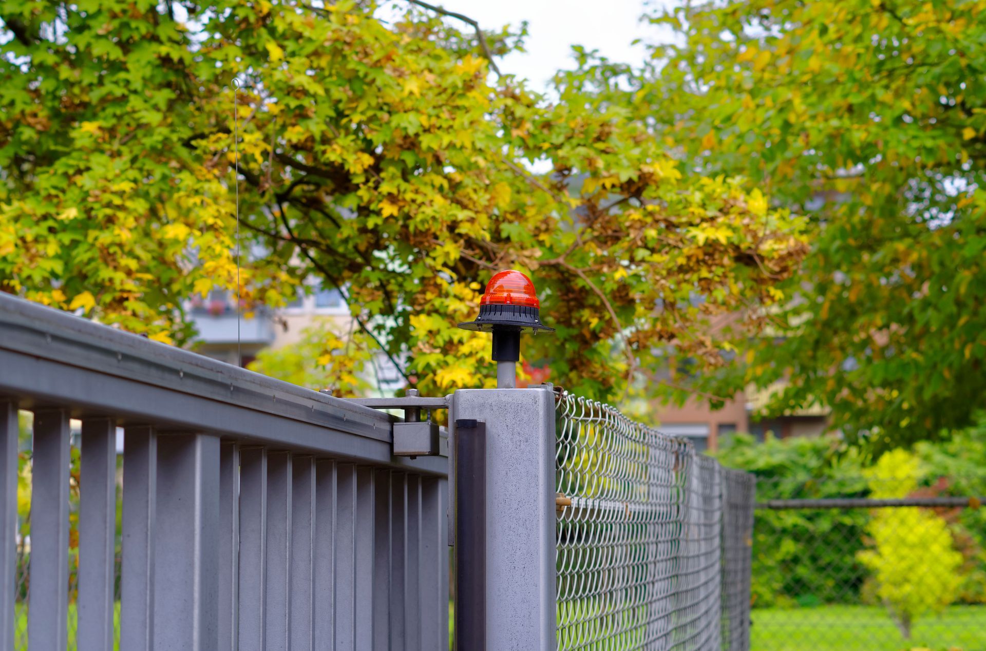 Metal fence with orange warning light, autumn trees in background.