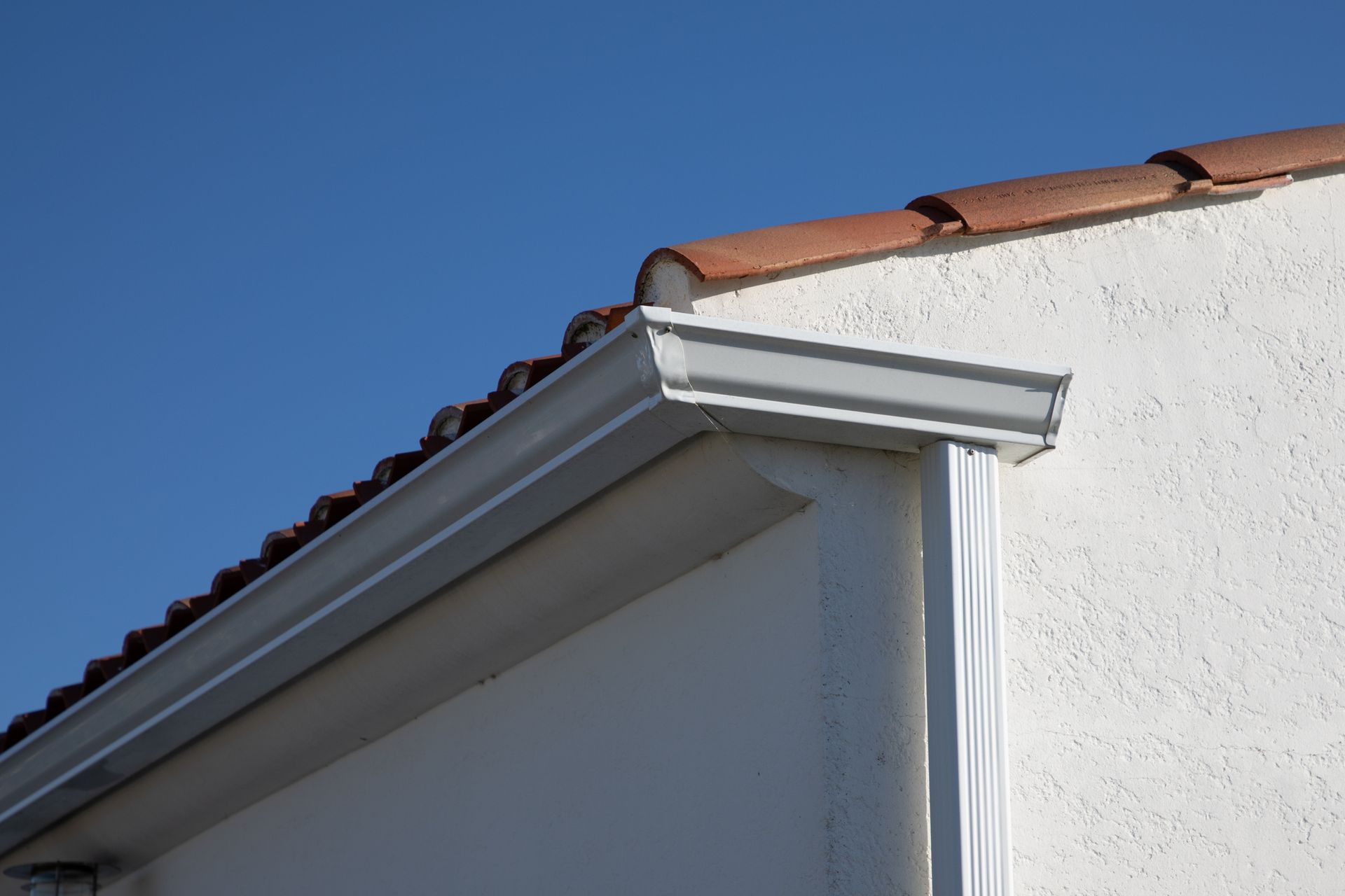 White gutter along a stucco wall under a terracotta tile roof, blue sky background.