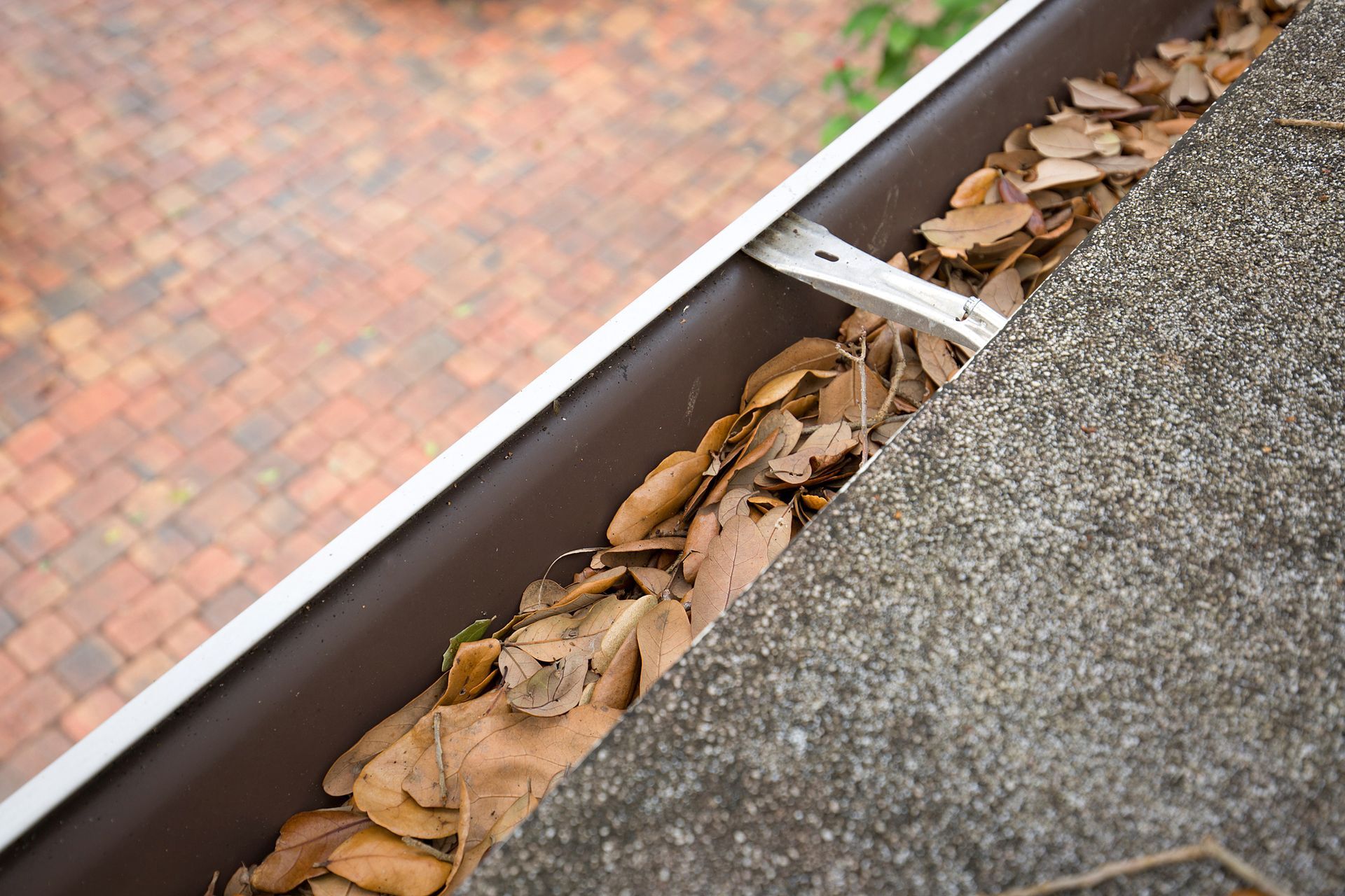 Brown gutter filled with dry leaves on a concrete roof next to a brick patio.