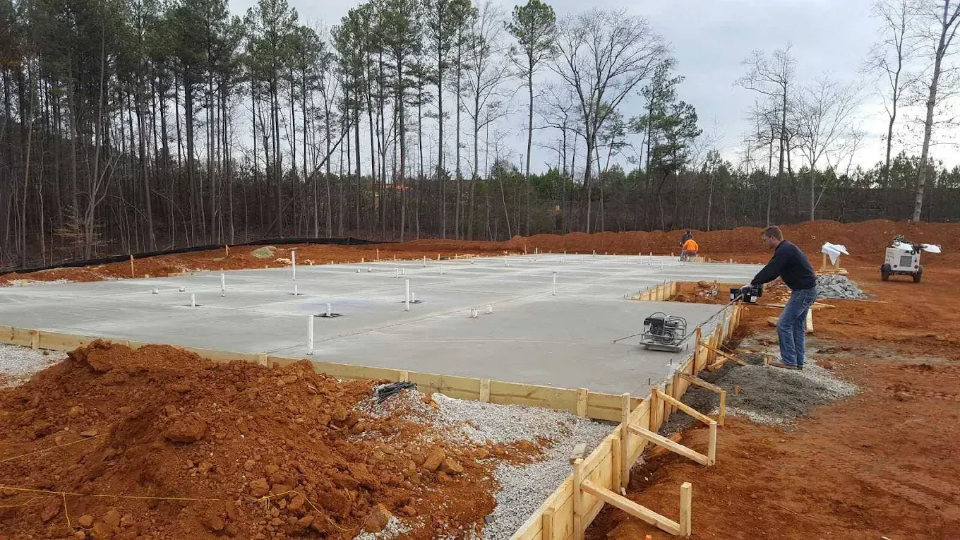 Construction site with a concrete foundation being worked on. A man operates a tool near wooden framing.