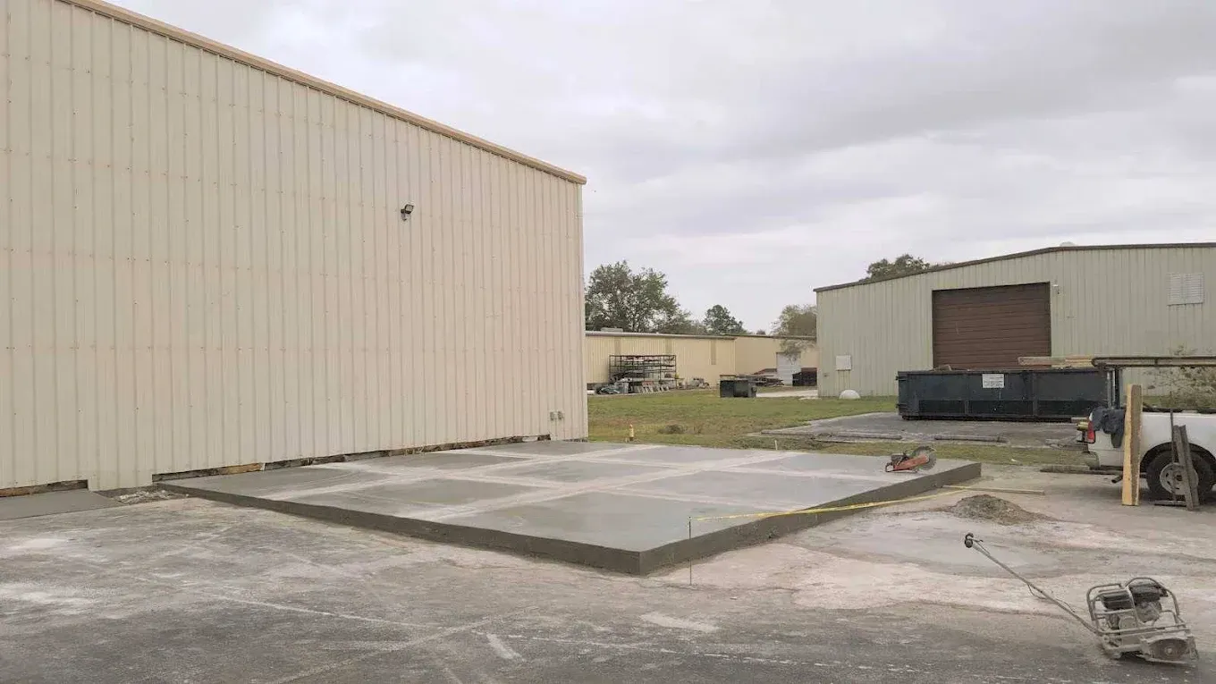 Concrete slab outside a beige industrial building, with another building and overcast sky in the background.