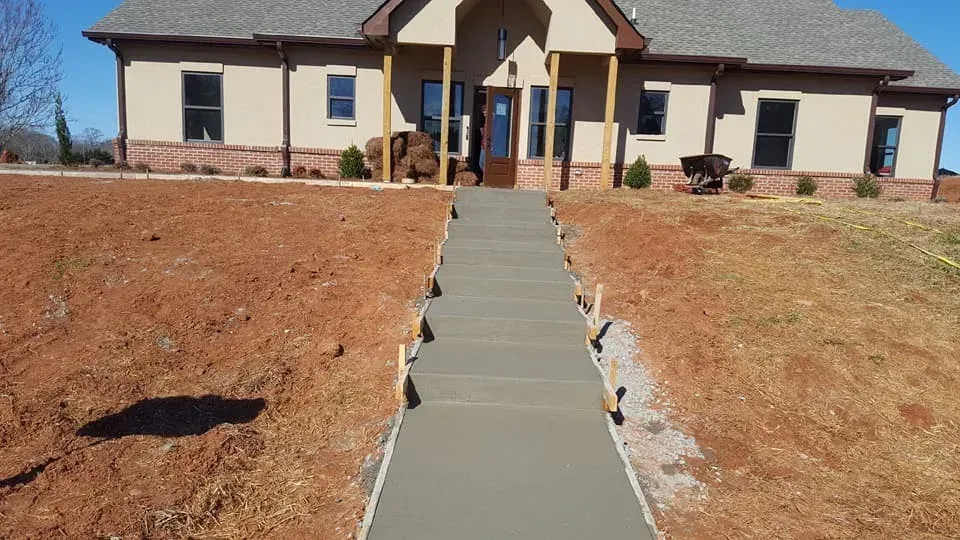 Newly poured concrete walkway leading to a beige house with brick trim on a sunny day.