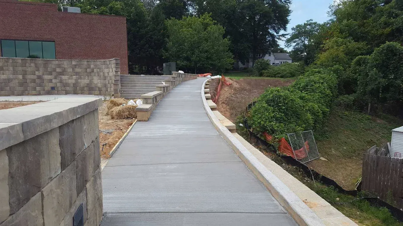 A concrete walkway with retaining walls winds between a brick building and overgrown greenery on a sunny day.
