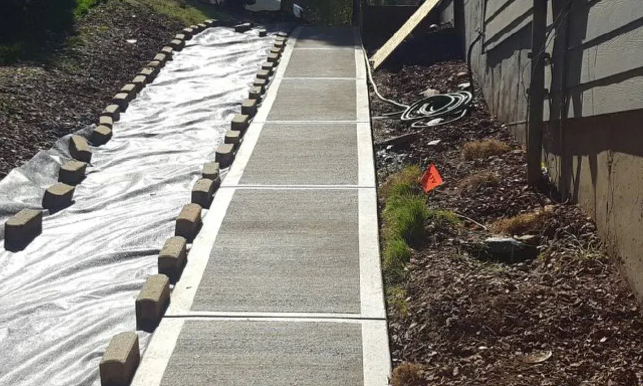 Concrete walkway with brick borders and landscaping, next to a house.