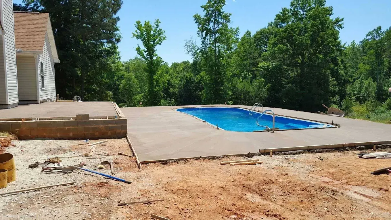Concrete pool deck under construction next to a house, with a blue pool and trees in the background.