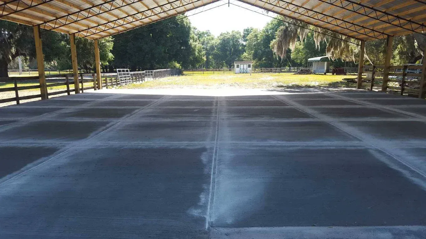 A concrete open-air building with a grid floor. The background shows grass, trees, and other structures.