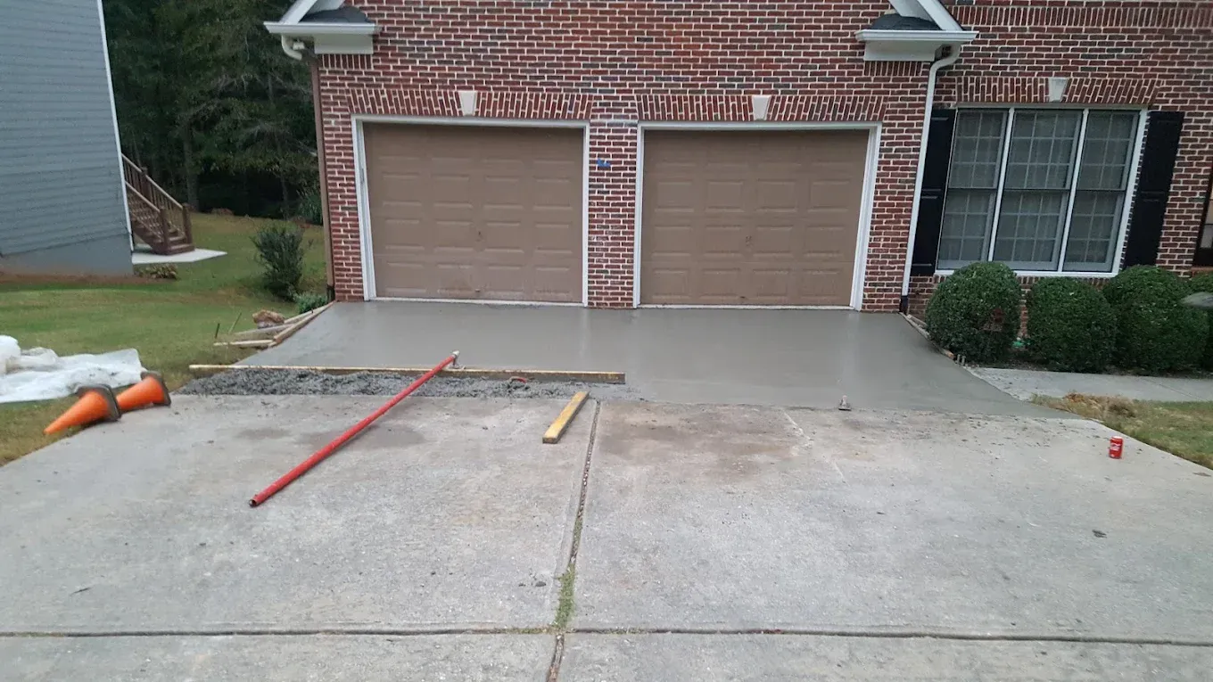 Newly poured concrete driveway in front of a brick house with garage doors. Tools and a traffic cone are visible.