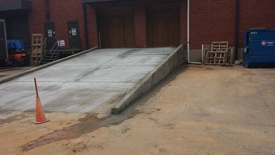 Concrete loading dock ramp at a brick building. An orange traffic cone sits near the ramp, and there is a dumpster and pallets nearby.