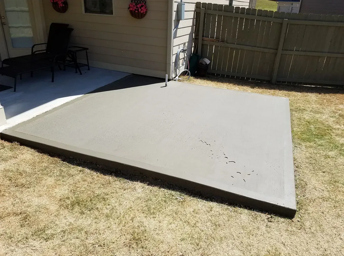 A freshly poured concrete patio in a backyard, next to a house and a fence.