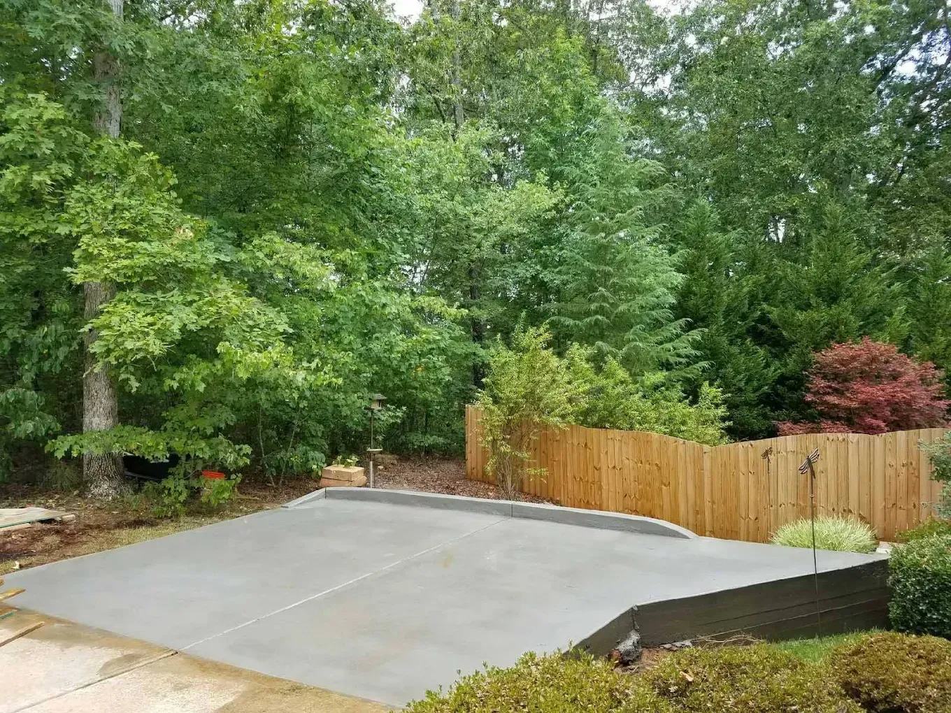 Concrete patio in a backyard, surrounded by a wooden fence and lush green trees.