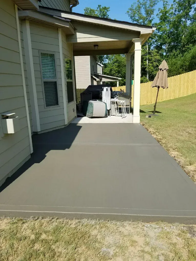 Newly poured concrete patio next to a house, with covered area and outdoor grill. Sunny day.