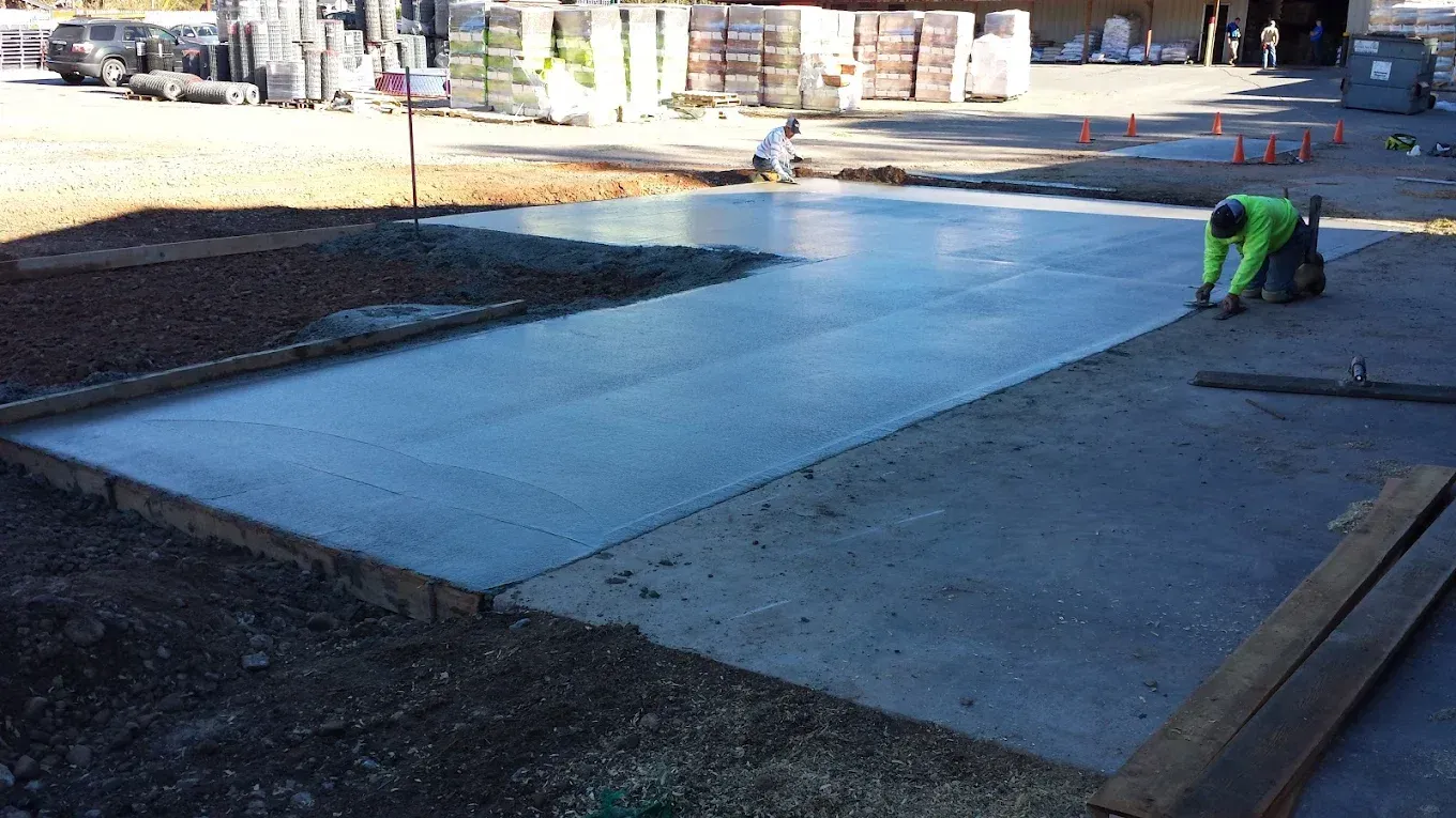 Construction site: worker smoothing wet concrete, rectangular pad. Sunlight, brown earth, supplies in the background.