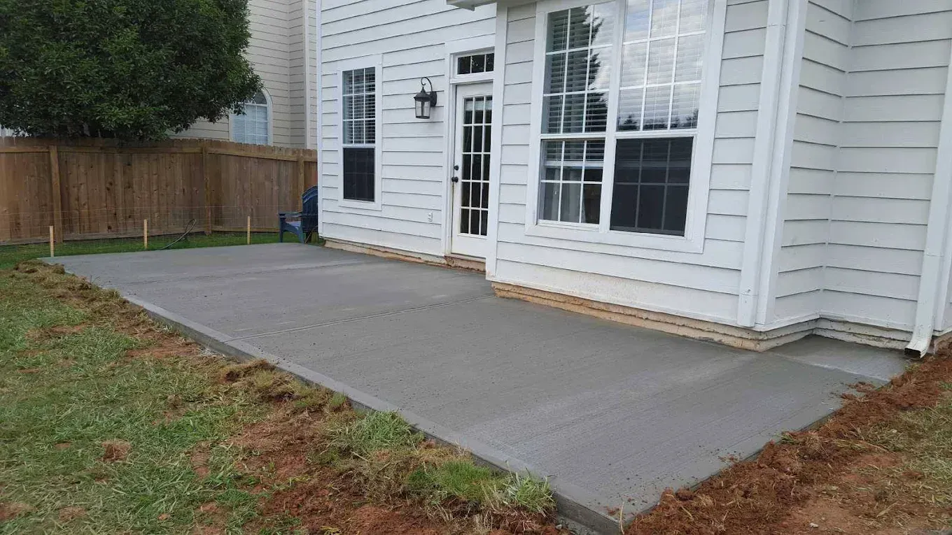 Newly poured concrete patio next to a white house with large windows. Lawn and a fence are in the background.