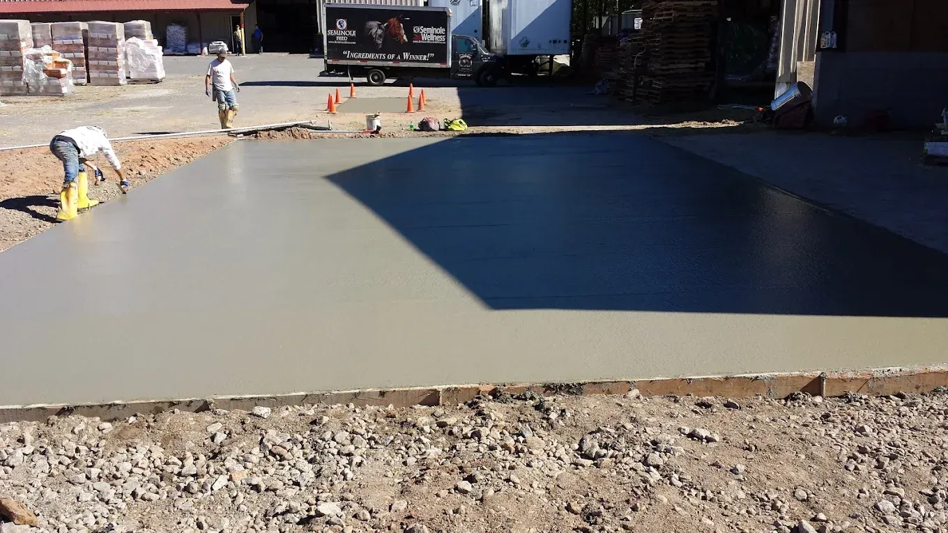 Workers leveling wet concrete on a construction site, near a truck and building.