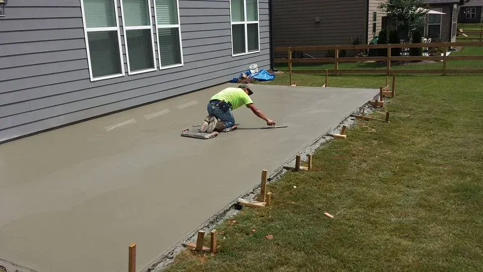 Man finishing a concrete patio next to a house. Green shirt, gray concrete, sunny day.