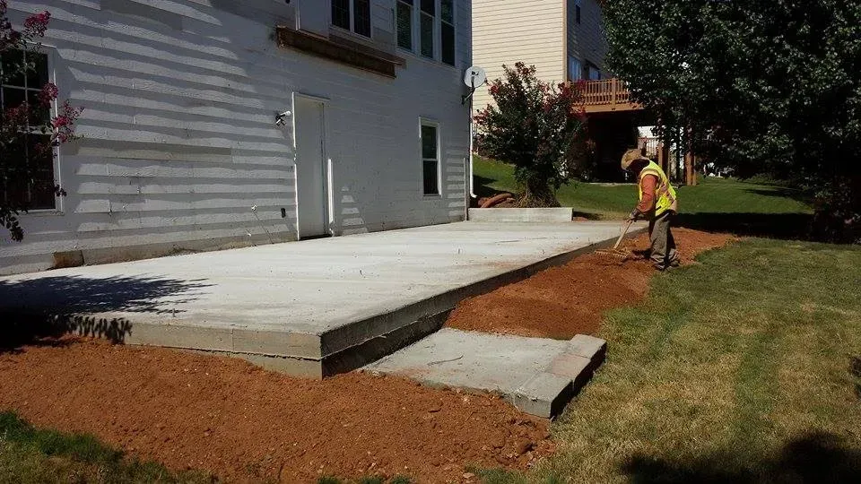 A person rakes brown soil near a concrete patio and house. Green grass, blue sky.