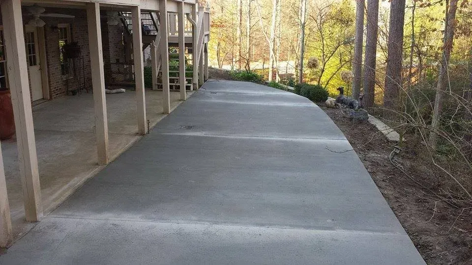 A long, newly poured concrete walkway beside a wooden deck, curving towards a wooded area.