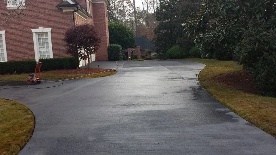 A wet asphalt driveway curves towards a brick house, bordered by grassy lawns and trees.