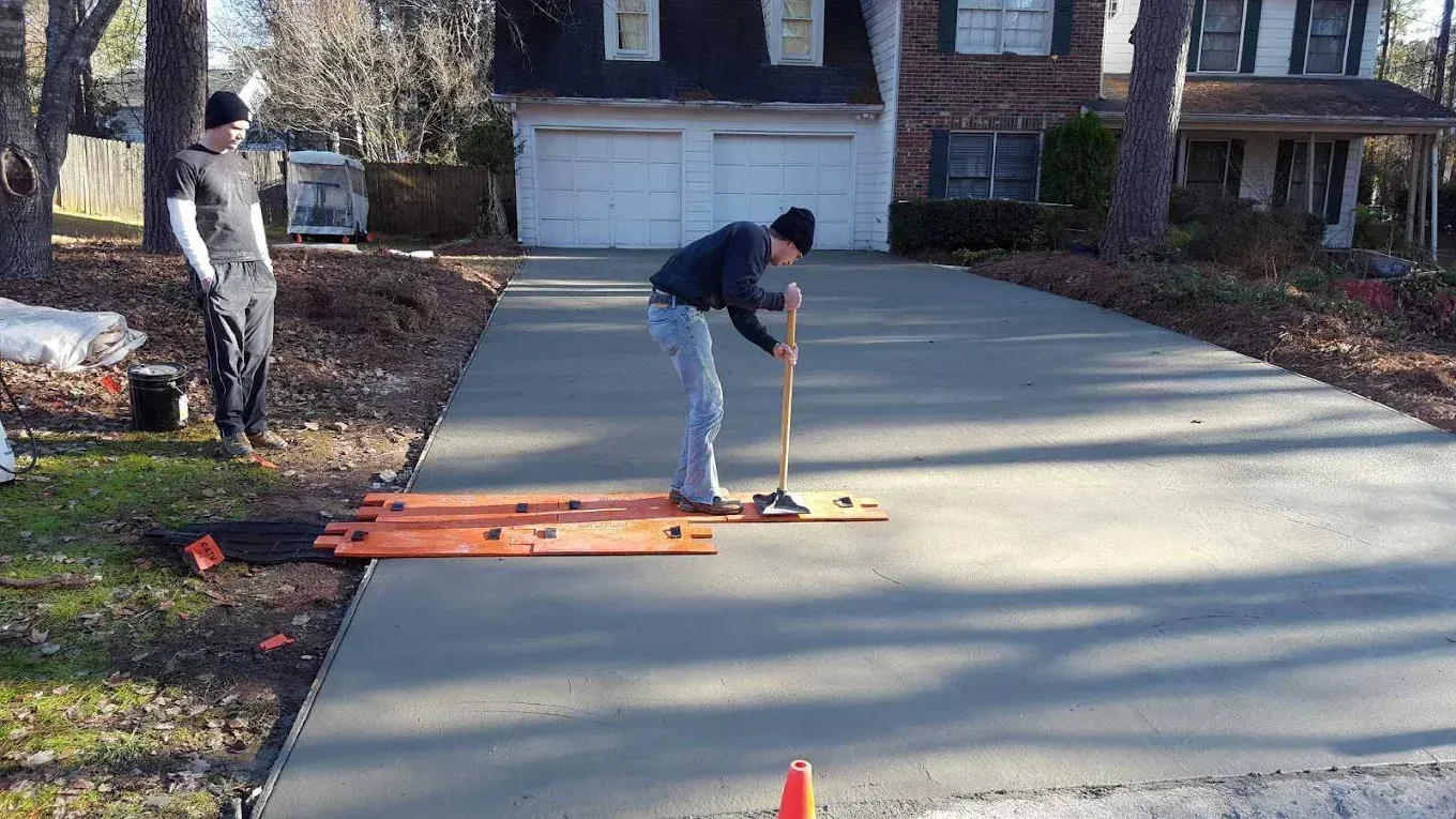 Two men working on a new concrete driveway. One uses a brush. House and trees in the background.