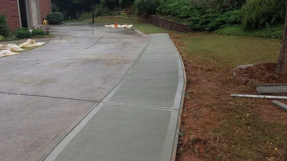 A concrete driveway transitions to a newly poured sidewalk, alongside grass and trees.