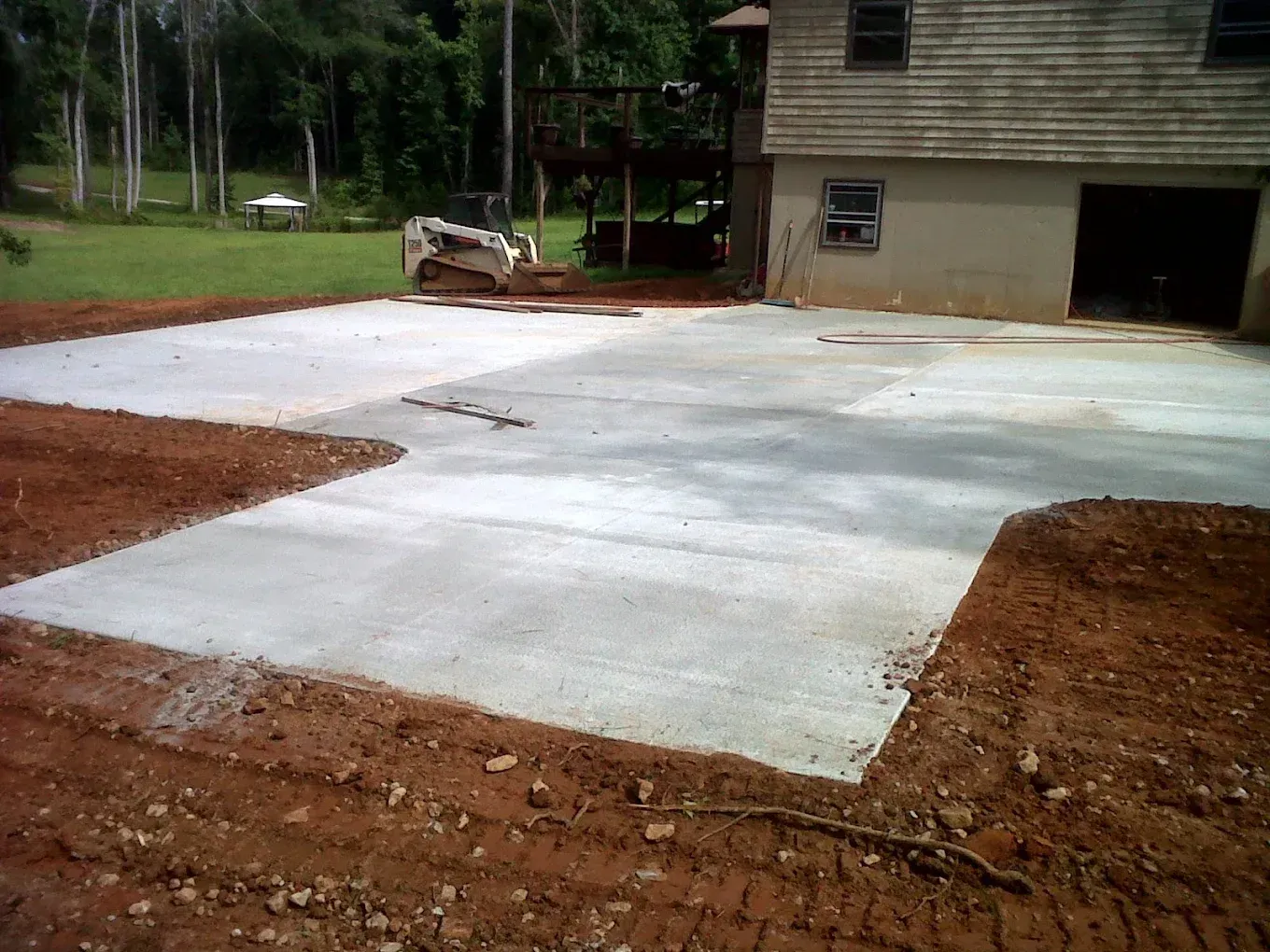 Newly poured concrete driveway next to a house. Brown earth surrounds the new surface; a small excavator sits in the background.