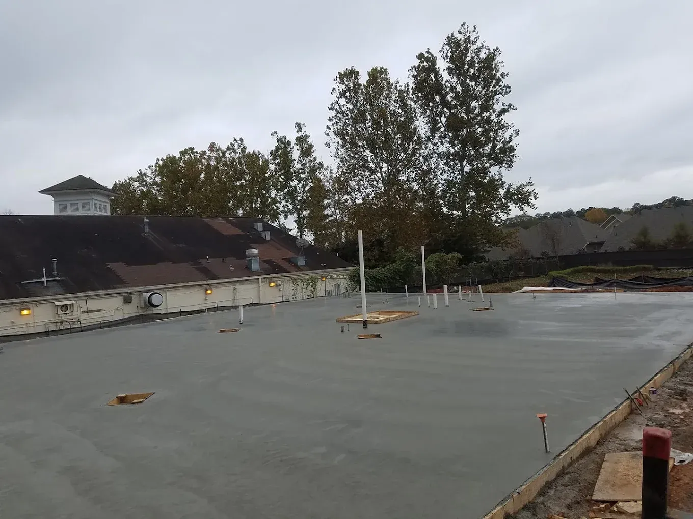 Newly poured gray concrete slab with pipes extending, a building and trees in the background.