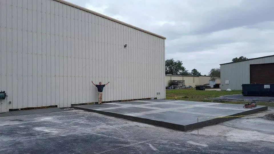 Man standing with arms up in front of a concrete slab next to a white industrial building under a cloudy sky.