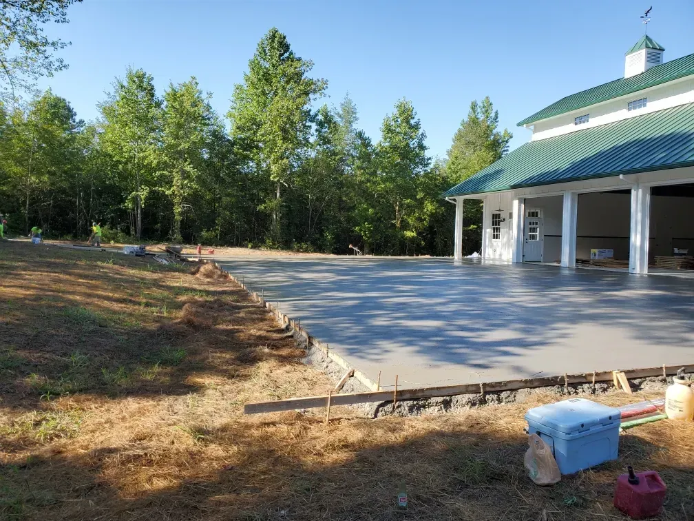 Newly poured concrete patio next to a white building with a green roof; trees in the background.
