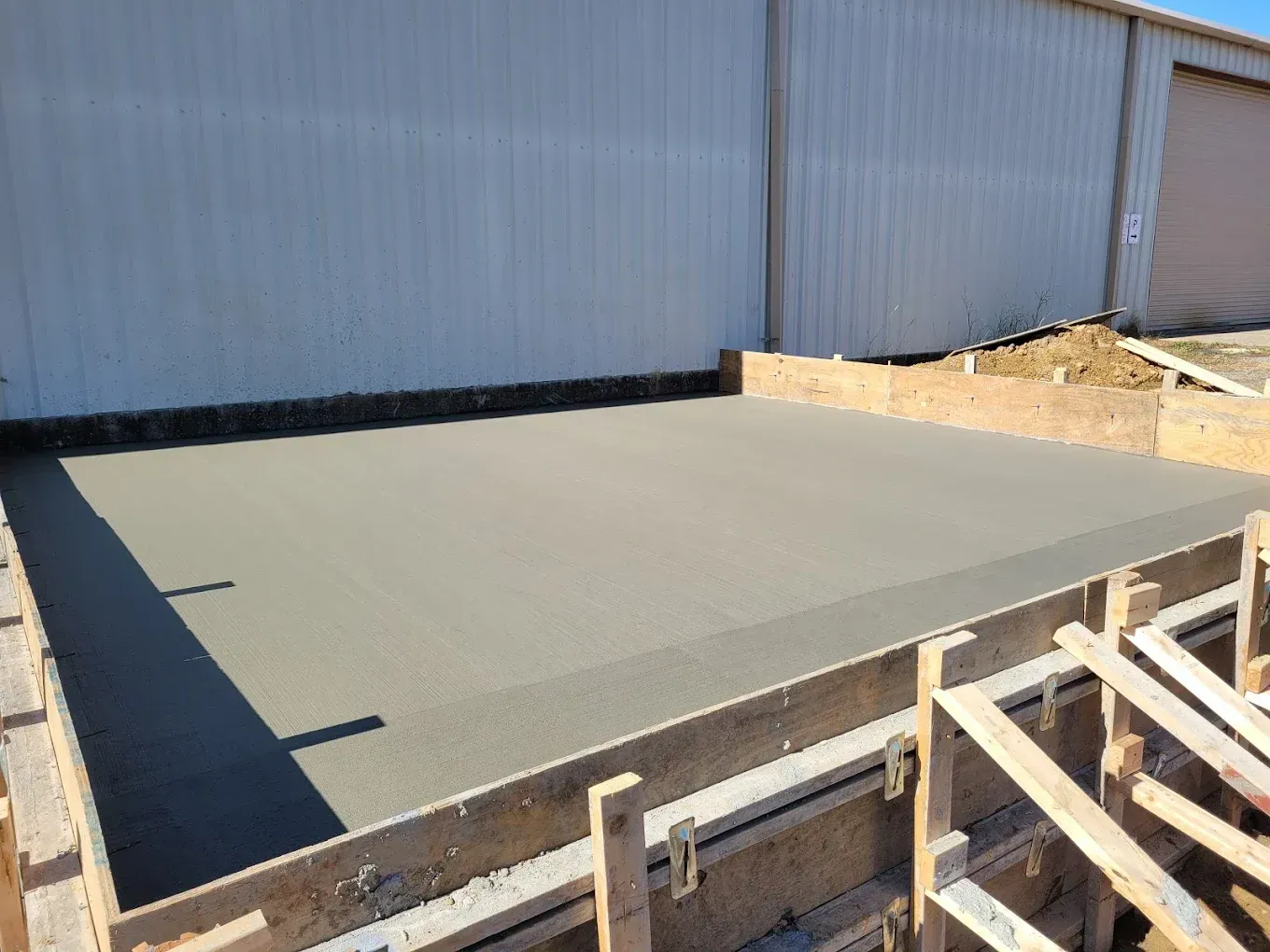 Freshly poured concrete slab within wooden forms, adjacent to a metal-sided building.