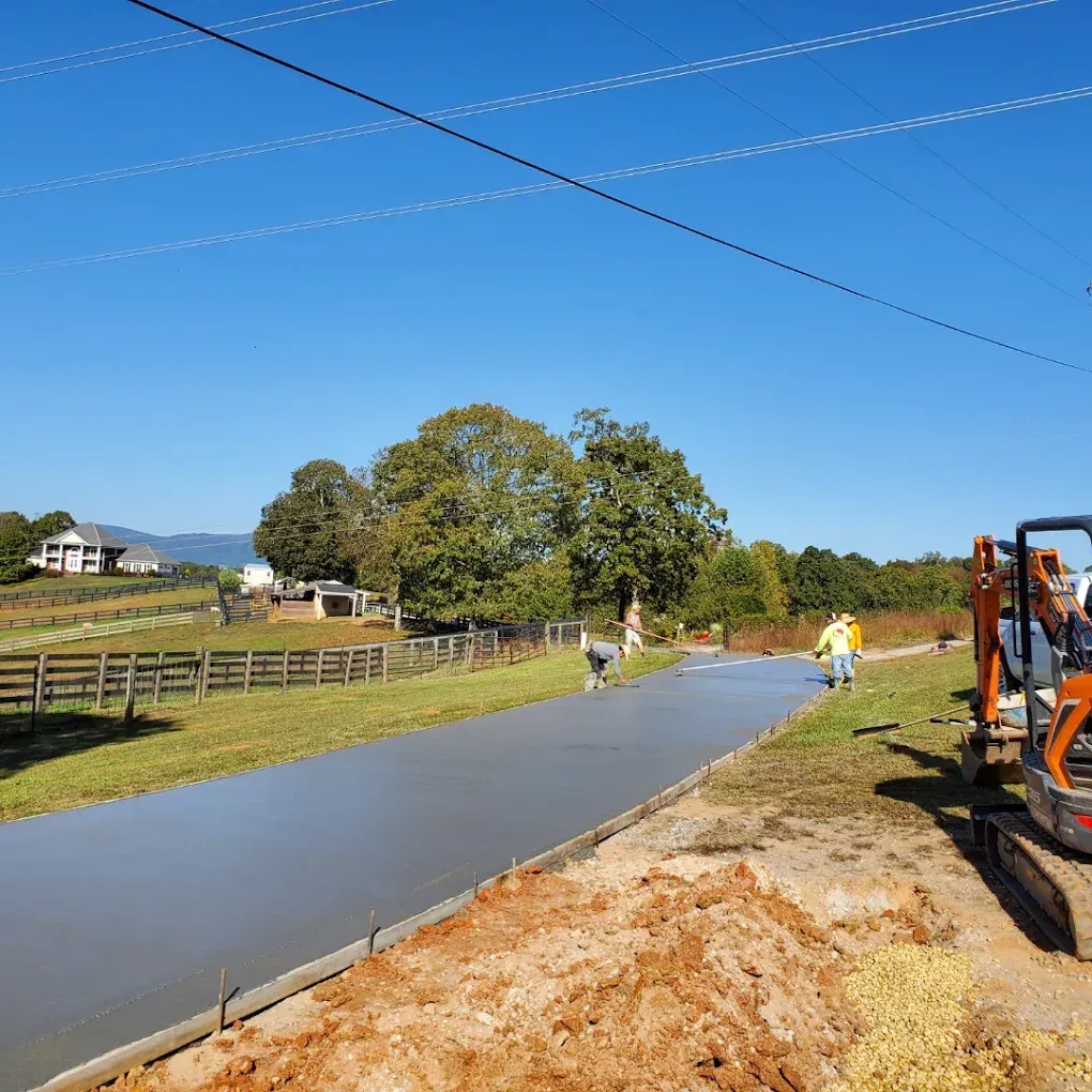 Workers smoothing fresh concrete on a path under a blue sky, with a tractor and rural scenery.