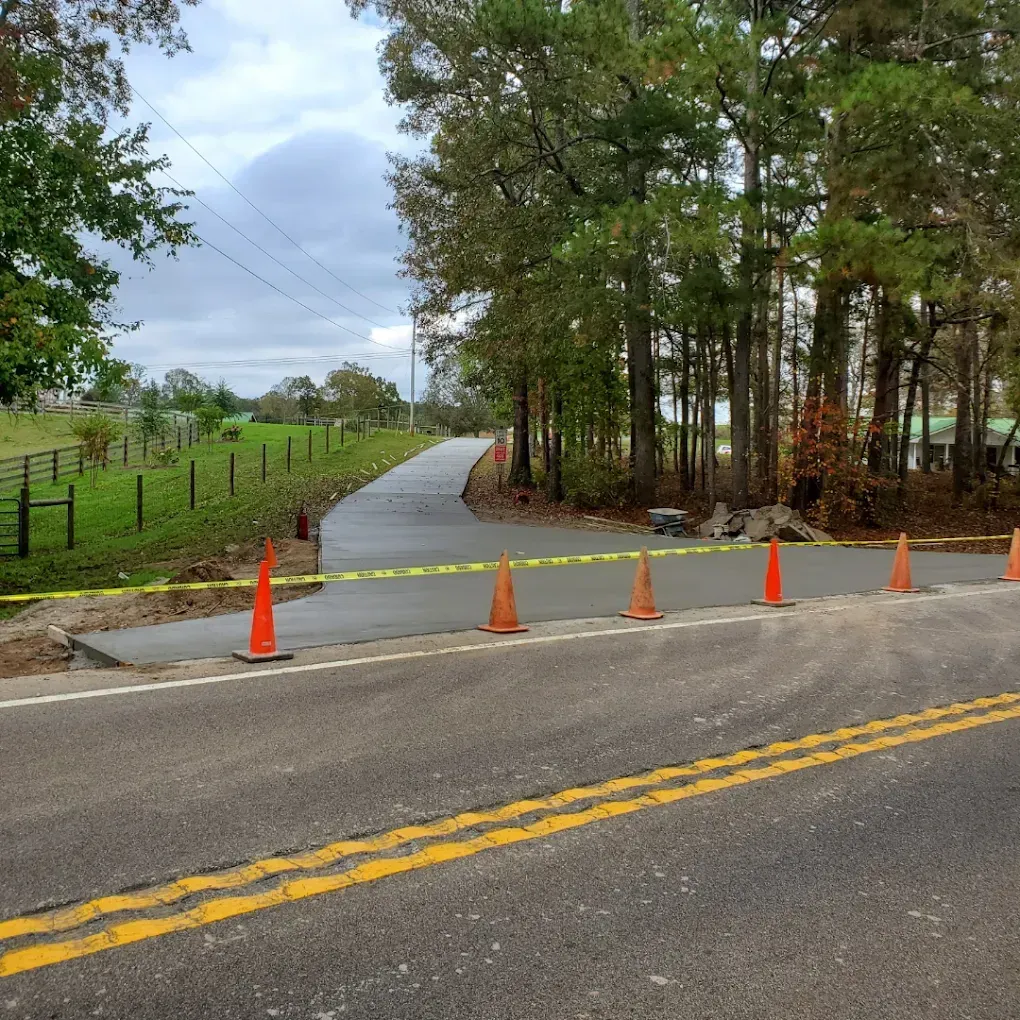 Road construction: freshly poured concrete driveway section blocked by orange cones and caution tape.
