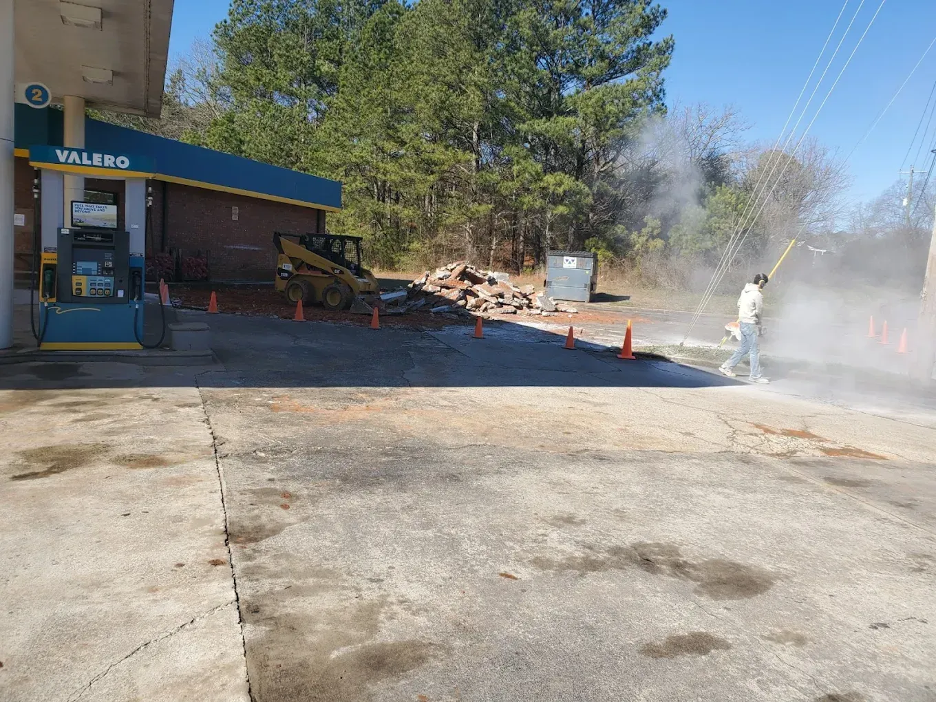 Person power washing a gas station's concrete. A skid steer is parked near stacked bricks with trees in the background.