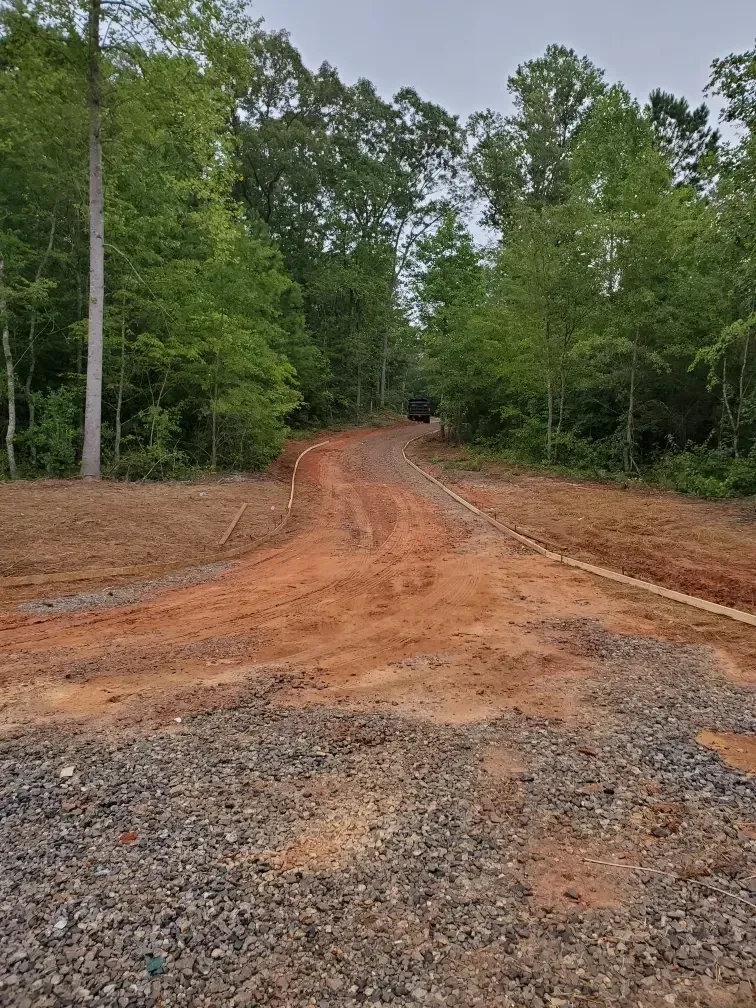 Dirt road through a wooded area with gravel, red clay, and trees. Cloudy sky.