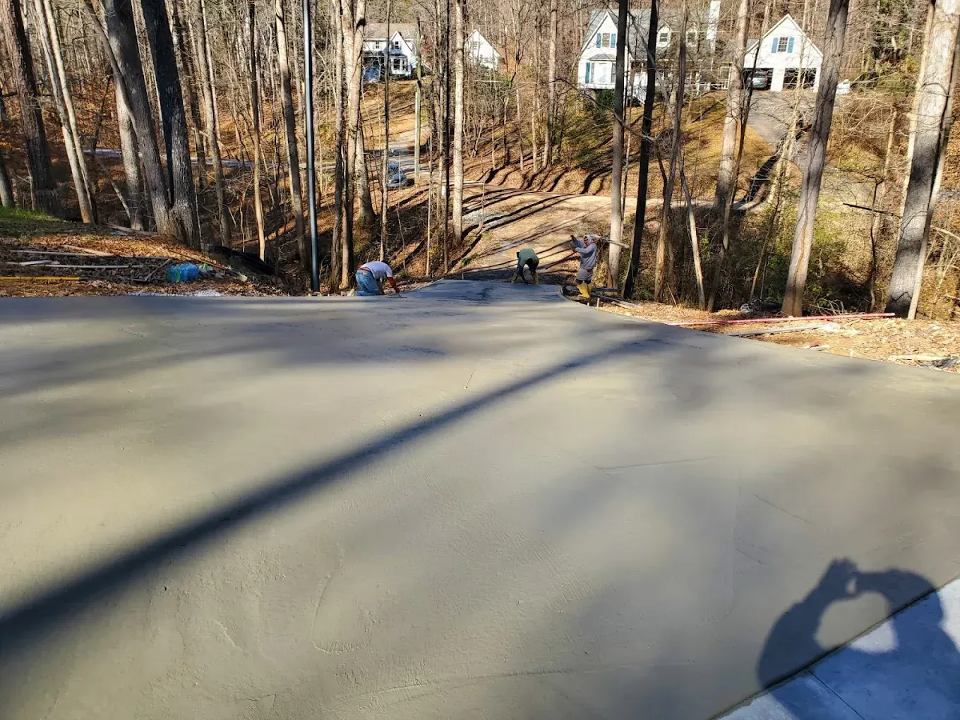 Concrete driveway being poured on a hillside. Workers in the distance, trees line the side, houses visible.