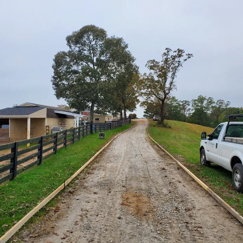 Dirt driveway bordered by wooden rails; a white truck parked on the right; building on the left.