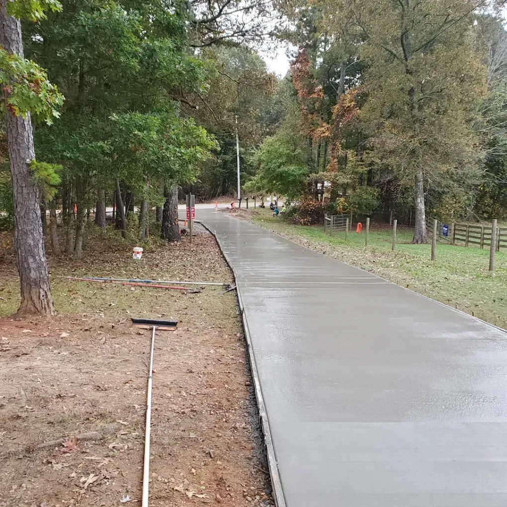 Newly poured concrete driveway in a wooded area. A rake lays nearby.