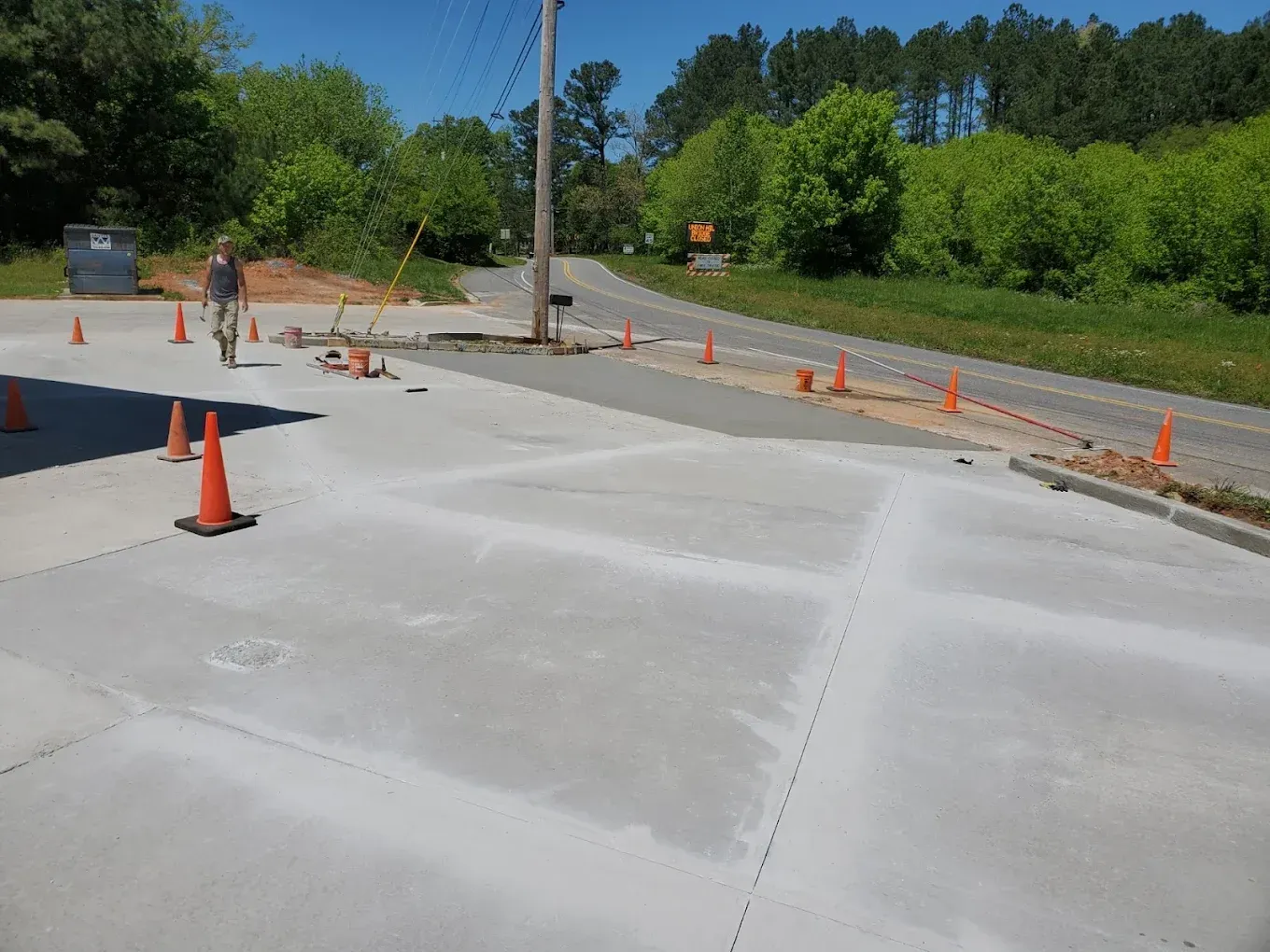 A person walks across a newly paved concrete surface with orange cones. A road and trees are in the background.