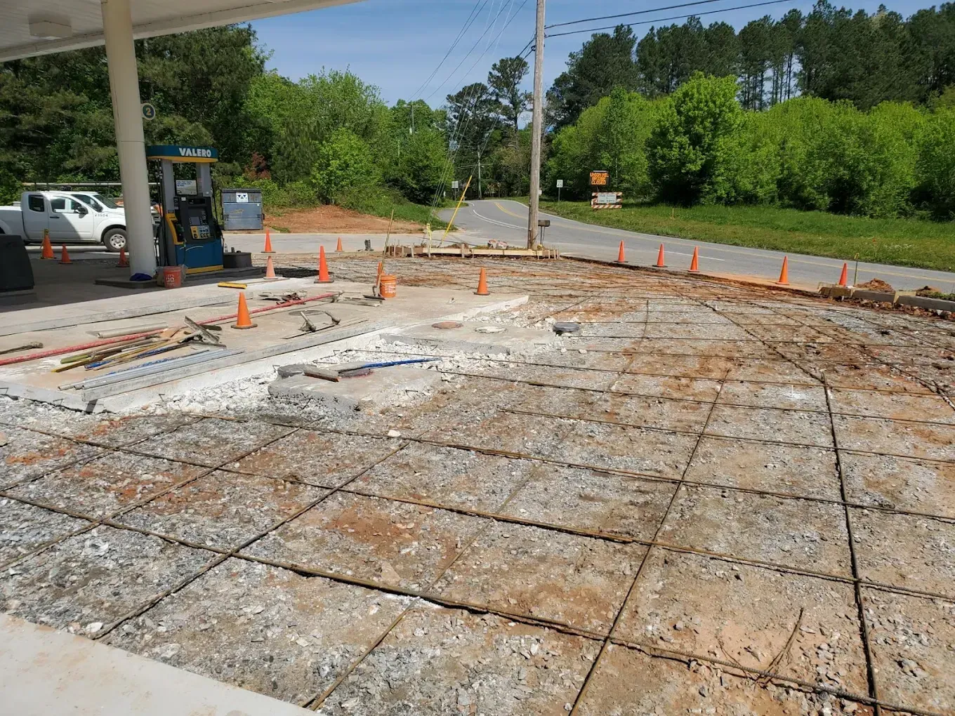 Gas station construction: concrete rebar grid, orange cones, vehicles, trees, blue sky.
