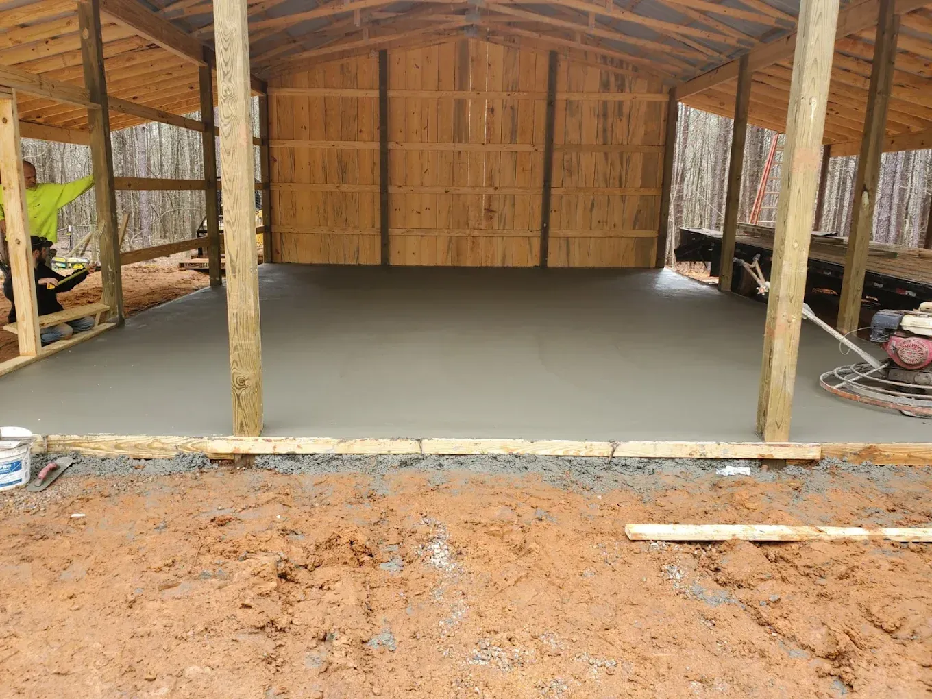 Newly poured concrete floor inside a wooden structure under construction, surrounded by dirt and trees.
