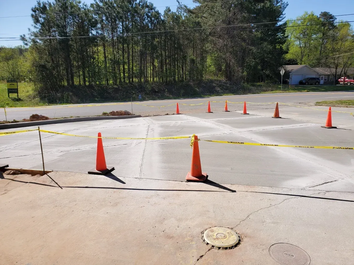 Freshly poured concrete area with orange cones, yellow caution tape, and trees in the background.