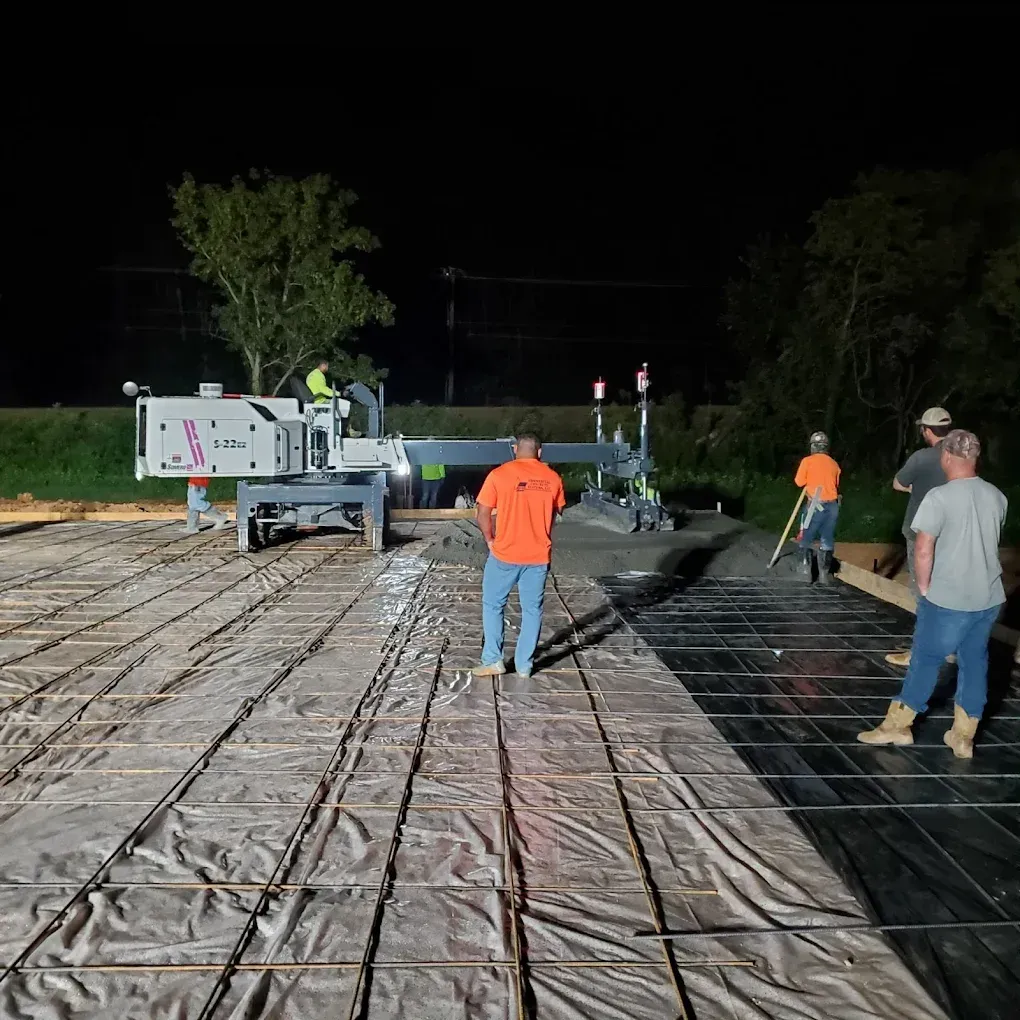 Construction workers pouring concrete at night; machine, rebar, and a level.