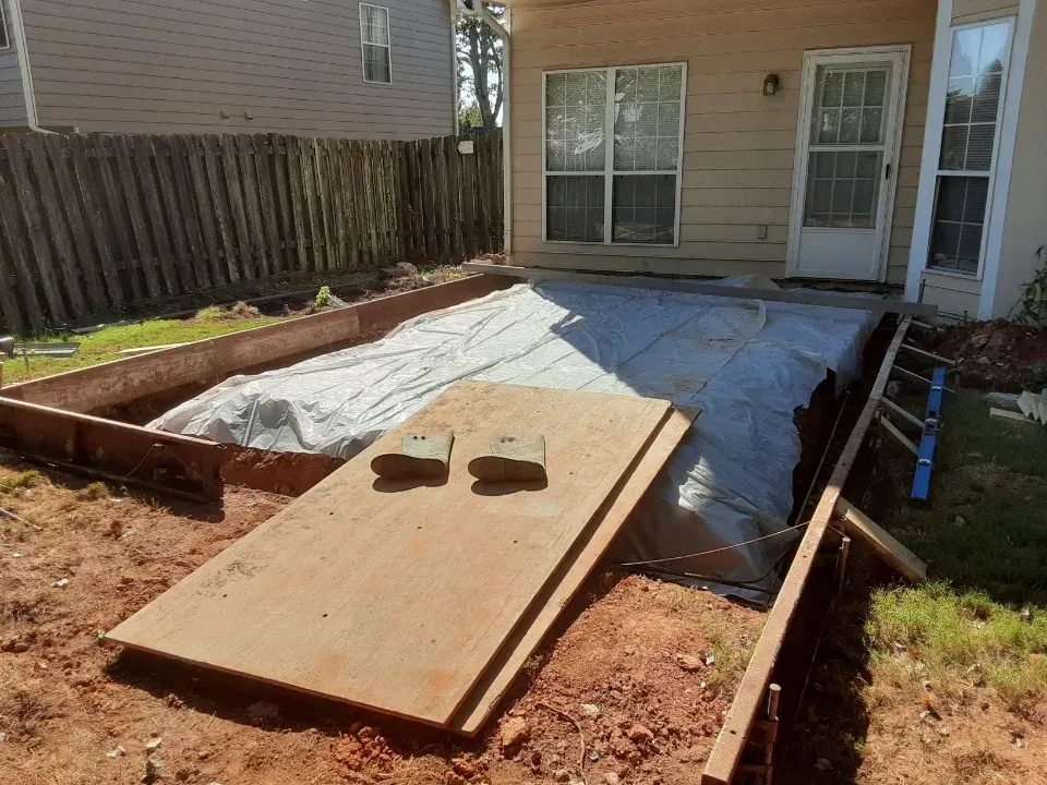 Construction site: wooden frame for patio, covered with tarp. Brown dirt, concrete patio, and house in the background.