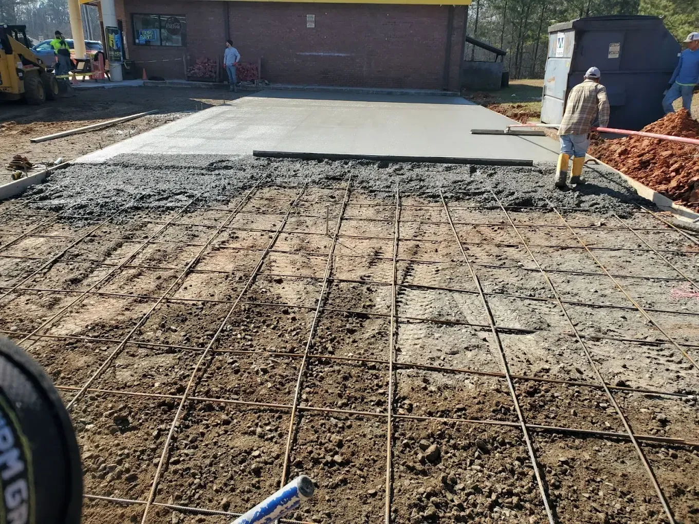 Construction workers pouring concrete over rebar on dirt ground. Yellow building in background.