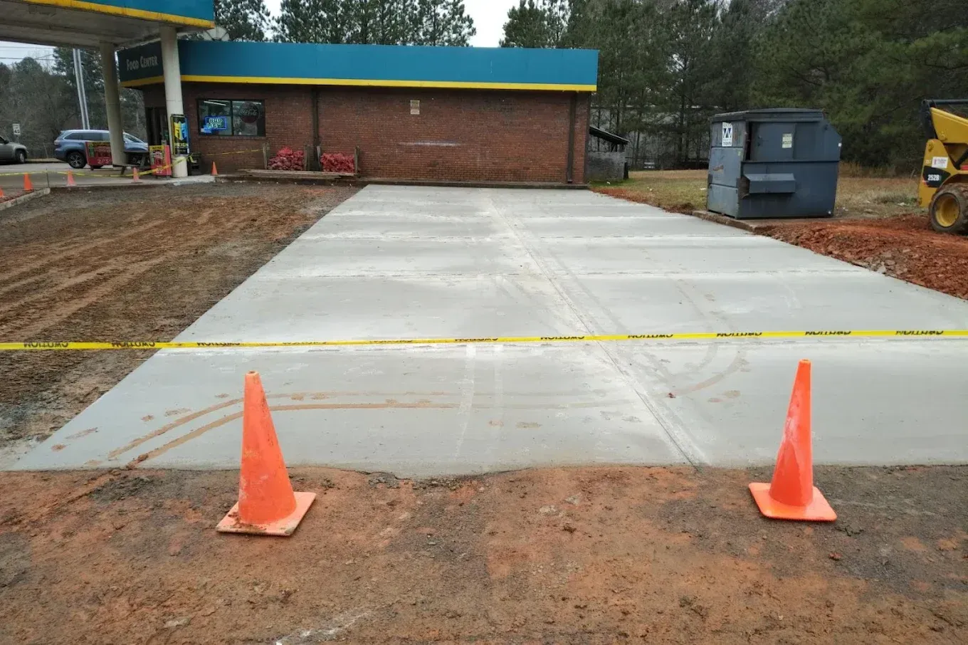 Orange cones mark a newly paved concrete area at a gas station, with caution tape, brick building, and machinery.