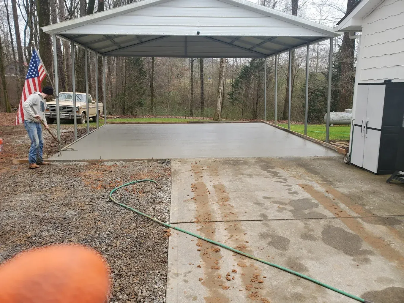 A person smoothing wet concrete under a metal carport; a car and house are visible in the background.