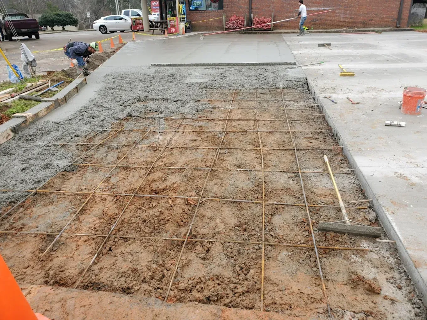 Construction site: worker pouring wet concrete over a metal grid. Other workers and vehicles visible.
