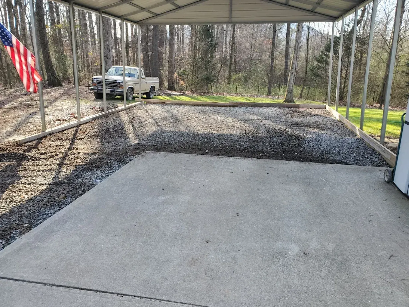 A concrete pad transitions to gravel under a carport, with a truck and American flag visible.